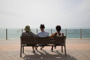 Three people sitting on a bench on the promenade look at the horizon of the Atlantic Ocean, photo taken from behind. People are on a trip in Cadiz, Spain. Concept travel and vacation.
