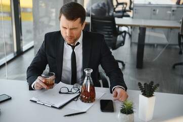 Fatigue young businessman sitting at workplace and holding whiskey bottle, drinking alcohol, bankruptcy concept.