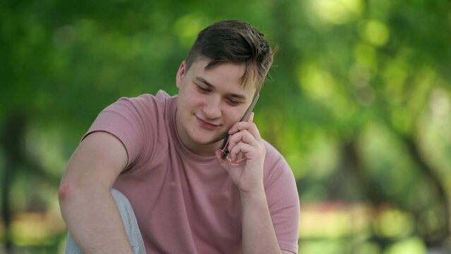 Medium shot portrait of positive Caucasian young man talking on phone sitting in sunny park. Front view confident guy chatting enjoying leisure on summer spring day outdoors