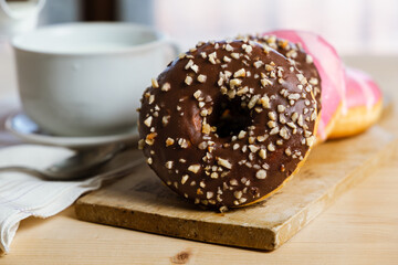 Close up view of donuts with chocolate and pink frosting over a wooden table, milk cup and milk poured, white towel in front of a window
