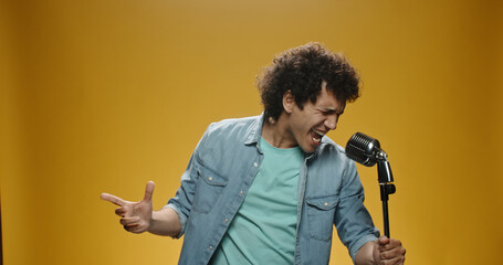 Close up shot of asian man with curly hair in casual clothes crazily dancing and singing in microphone, isolated on yellow background 
