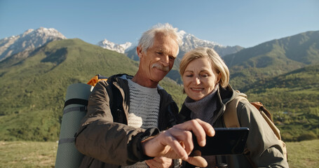 Old couple having a hike in spring mountains, then stopping to take a picture on a smartphone. Senior caucasian family spending time together travelling after retirement - tourism concept 