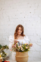 A young female florist in white clothes works against the background of a white brick wall, making bouquets in a flower shop. Workplace of a flower shop employee.