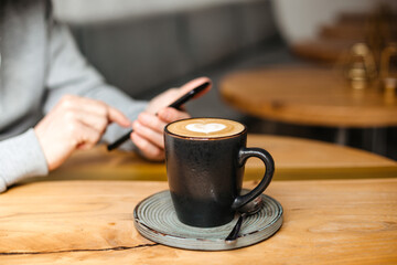 The man writes a message on his phone and drinks coffee. Cappuccino on vegetable milk in a black mug on a wooden table. Business meeting in a cafe