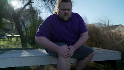 Person suffering from knee pain sitting at bench outdoors. One overweight caucasian male person having physical injury