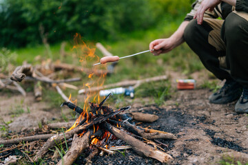 Closeup cropped shot of unrecognizable tourist male grilling sausages on campfire in countryside....