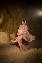 portrait of beautiful pregnant young brunette woman  in long vintage flying with wind dress holding her belly in desert studio in cave