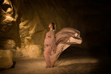 ortrait of beautiful pregnant young brunette woman in long vintage flying with wind dress holding her belly in desert studio in cave