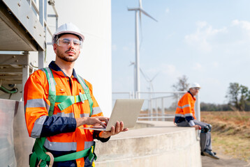 Caucasian engineer or technician hold laptop and look at camera also stay near base of windmill or wind turbine with his co-worker sit in th back.