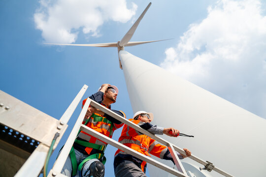 Two engineers or technician workers stand on base of big windmill or wind turbine and look to right side with blue sky.
