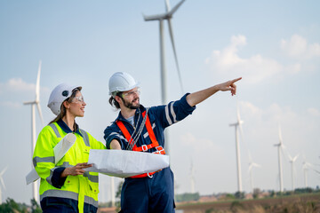 Caucasian engineer man point to right side and discuss together with co-worker woman hold drawing paper and stay in front of row of windmill or wind turbine with blue sky.