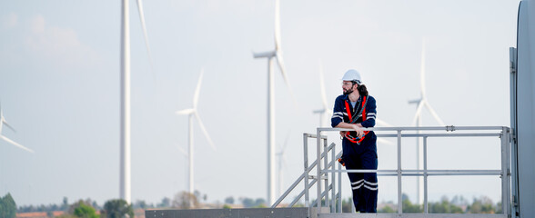 Wide horizontal image of engineer man or technician worker stand on base of windmill or wind turbine and look to left side also stay in front of windmill cluster in background with blue sky.