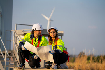 Engineer man and woman sit on the ground and discuss together with using drawing paper and stay in front of row of windmill or wind turbine base with blue sky and warm evening light.