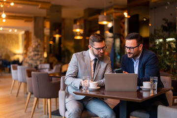 Two businessmen, professional managers using smartphone and laptop computer in co-working, checking corporate apps while sitting in city coffee shop or restaurant. Stylish men on meeting in coffee bar