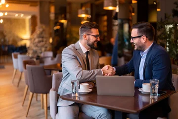 Keuken spatwand met foto Two elegant stylish corporate leaders shaking hands after a successful business meeting in a restaurant in the evening. Proud business partners shaking hands while sitting at table in restaurant. © Dorde
