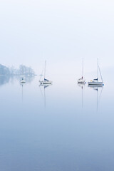 Calm lake with mirror-like reflections and yachts on a misty morning. Windermere, Lake District, UK. Peaceful British scenic views in the countryside. - 572016386
