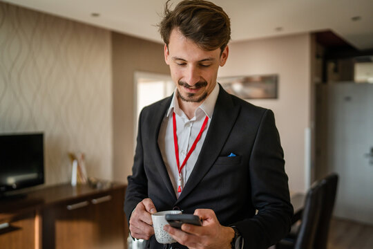 one man adult caucasian businessman wear suit in hotel room while taking a brake or prepare for work hold cup of coffee and smartphone sending sms messages copy space