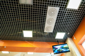 Various lighting fixtures and air conditioning grilles on the ceiling of the restaurant. LED lamps on the suspended mesh ceiling. Industrial air conditioning in the large hall.