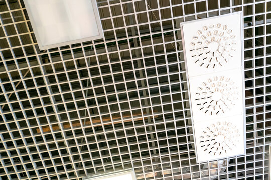 Various lighting fixtures and air conditioning grilles on the ceiling of the restaurant. LED lamps on the suspended mesh ceiling. Industrial air conditioning in the large hall. - Powered by Adobe