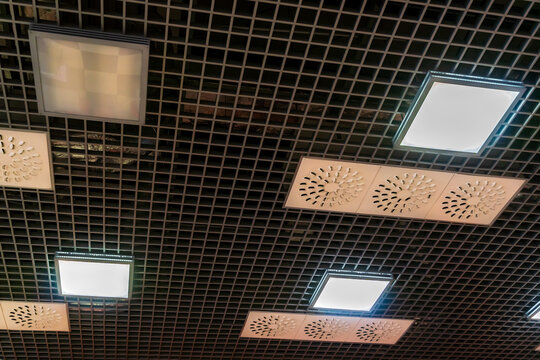 Various lighting fixtures and air conditioning grilles on the ceiling of the restaurant. LED lamps on the suspended mesh ceiling. Industrial air conditioning in the large hall. - Powered by Adobe
