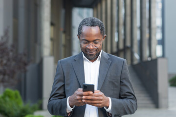 An African American businessman in a suit and glasses stands outside near an office center and uses a telephone. Writes messages, reads news, checks mail.