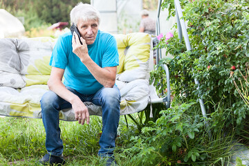 Fototapeta premium Elderly man sitting on a hanging chair and talking on the phone in the country house backyard