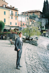 Fototapeta na wymiar Man in a suit stands on a paved embankment with houses in the background. Varenna, Italy