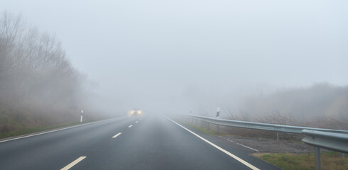 Auto fährt im Nebel auf einer Straße. Die Scheinwerfer leuchten hell. 