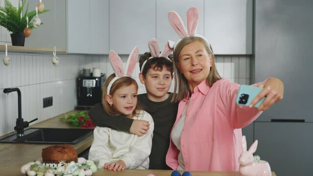 Smiling grandmother and grandchildren doing selfie with easter eggs, wearing bunny ears, have beauty and fun day together at home. Happy easter. Concept of childhood, technology, family weekend