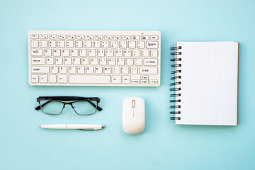 Office workspace with keyboard, notepad, glasses, pen and coffee cup. Flat lay image on blue, minimal.