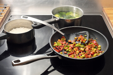 Diced vegetables are stir-fried with spices in a frying pan, pots with rice and broccoli behind them on the black stovetop, Asian style cooking, copy space, selected focus