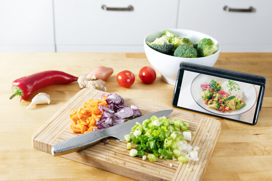 Cut vegetables on a wooden kitchen board, picture of the finished recipe on the smartphone, cooking by using an online app, modern preparation of a healthy meal, selected focus