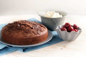 Chocolate cake and bowls with cherries and whipped cream on a blue napkin and a light wooden table, copy space, selected soft focus