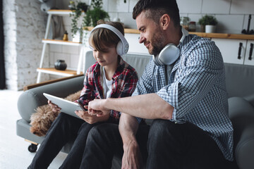 Handsome young strong father and his teenage son spending quality time together, having fun, enjoying togetherness. Boy playing tablet, learning, listen to music, dad helps