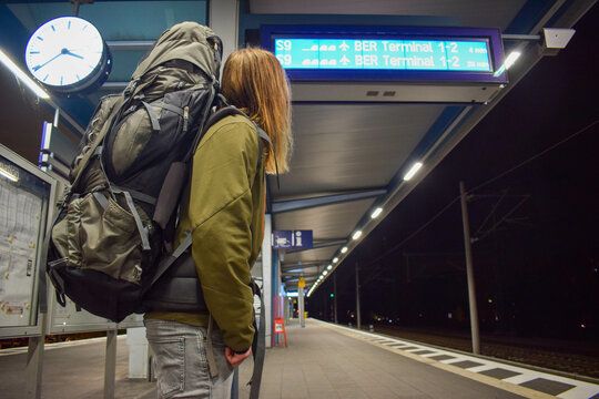 Young man with glasses and long hair standing inside train station looking at schedule alone in subway with big backpack on trip in urban city public transport while in adventure excursion vacation