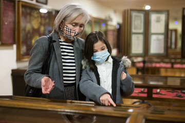 Positive tween schoolgirl and friendly elderly female tutor in face masks observing with interest arts and crafts on exhibition in art gallery. New normal in coronavirus pandemic