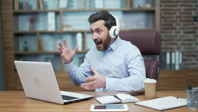 Excited mature bearded fan businessman with headphones looking at laptop computer screen watch online sport game cheering and celebrating win goal rejoices in victory good result at office workplace