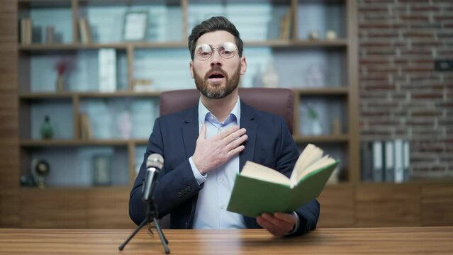 Webcam view of mature preacher pastor with glasses looking at the camera while reading the bible online talking into microphone during recording distance sermon podcast by video conference indoors