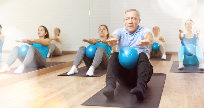 Willing aged man engaging in pilates training with ball in gym room during workout session. Persons practicing pilates in fitness studio