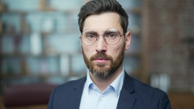 Close up portrait face of confident bearded businessman investor with glasses looking at the camera at modern office workplace Successful serious entrepreneur owner in formal suit indoors alone