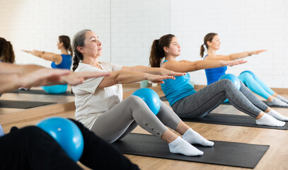 Fototapeta premium Sedulous old-aged woman doing pilates exercises with ball in gym area during workout session. Persons engaging in pilates in fitness room