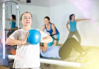 Fototapeta na wymiar Smiling old female pensioner squeezing ball in hands during Pilates training in rehabilitation center
