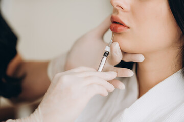 Close up of hands of cosmetologist making botox injection in female lips. She is holding syringe. The young beautiful woman is receiving procedure with enjoyment.