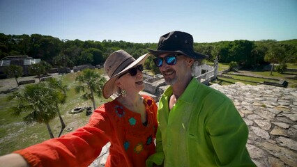 Mature woman and man couple wearing ethnic clothes, sunglasses, hats taking selfies at the top of Xcambo Mayan pyramid ruins in Mexico. - Powered by Adobe