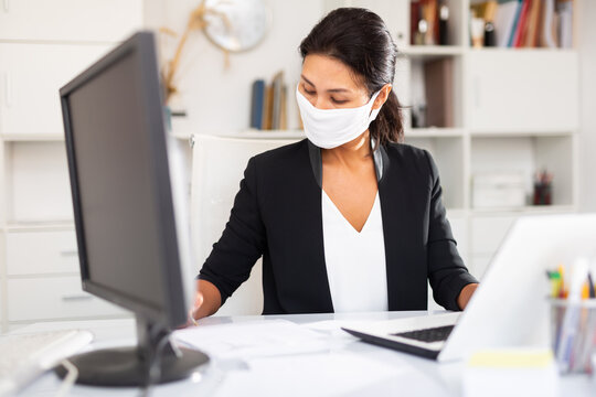 Professional business woman in protective medical mask using laptop at workplace in office