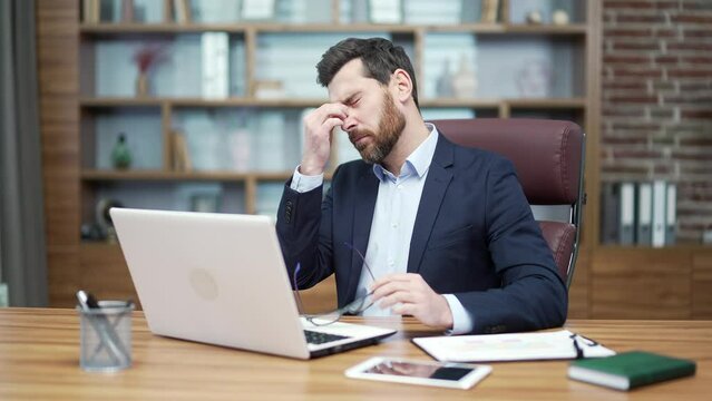 Tired overworked entrepreneur business man investor with eyes hurt massaging having bad poor vision after hard day working on laptop computer typing and browsing at desk in modern office indoors 