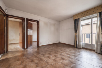 Empty room with old brown stoneware floor, wall air conditioner, cheap wooden access doors to other rooms and balcony with curtains and sheers