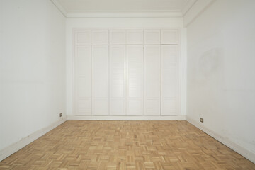 Empty room with one wall covered in white wooden cabinets mezzanine trunks all with white Venetian type doors, and checkerboard shaped oak slatted floors