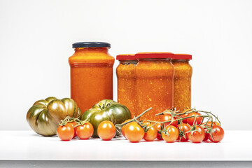 A still life with tomato sauce boats and pasta sauces with sweet marmalade tomatoes and bunches of cherry tomatoes on a white surface