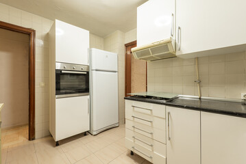 Corner of an old kitchen with white furniture, a column with an oven and a white fridge